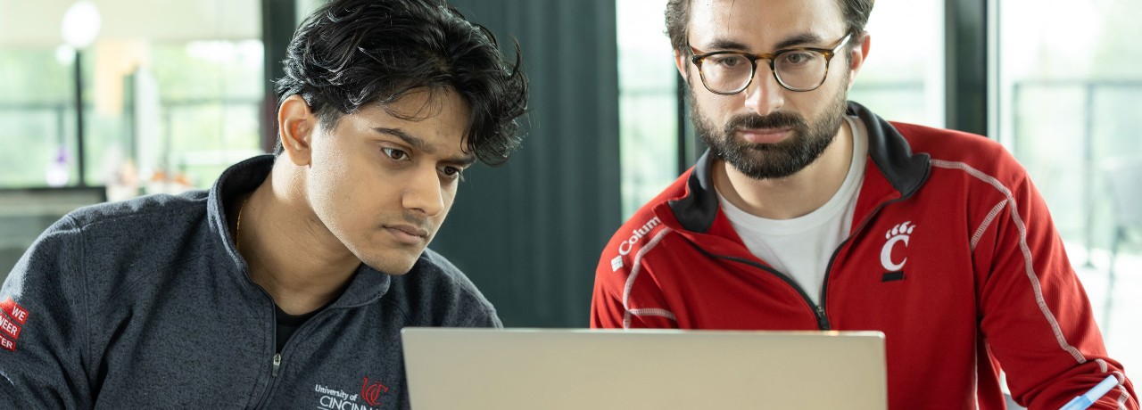 Two individuals in University of Cincinnati branded clothing are focused on a laptop screen in an office setting.