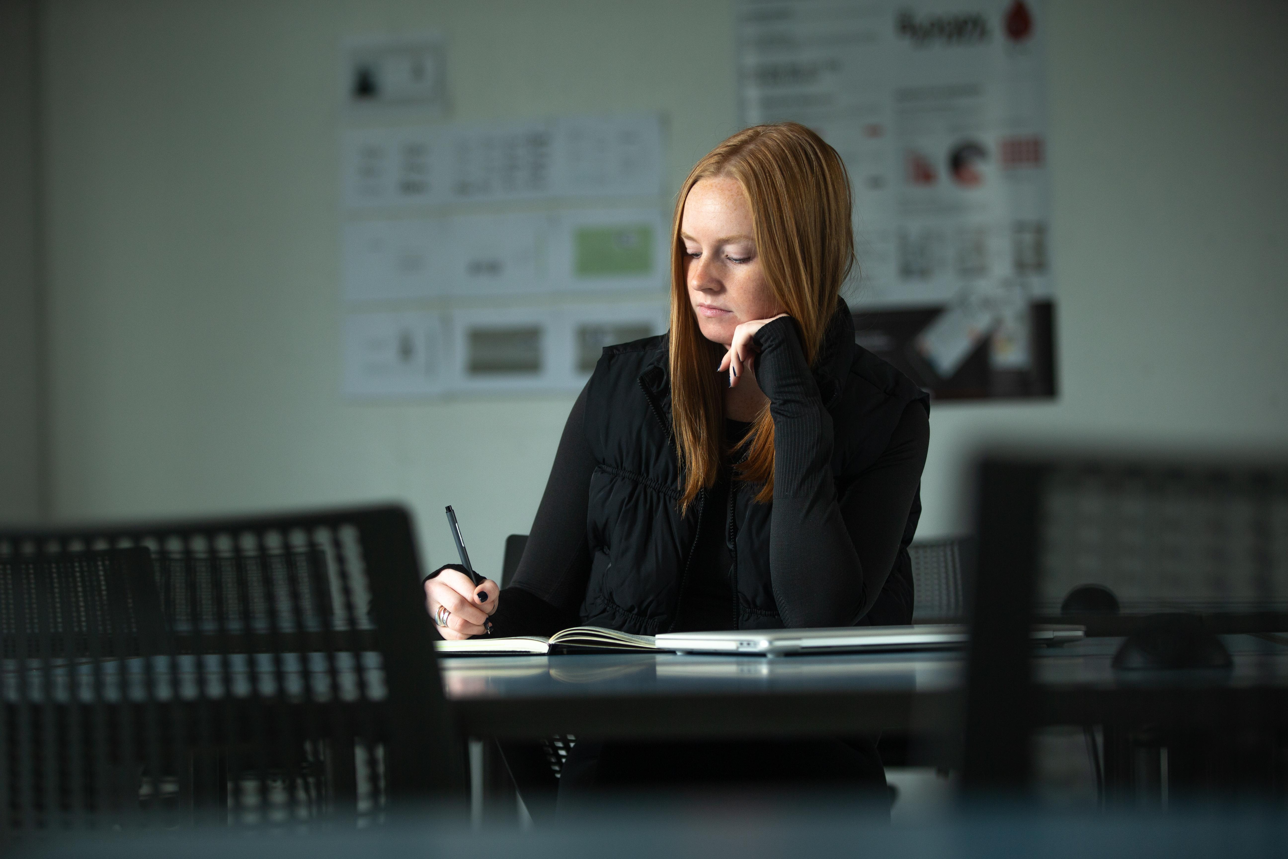 A student sits at a conference room table with pen in hand looking down at the paper while writing.