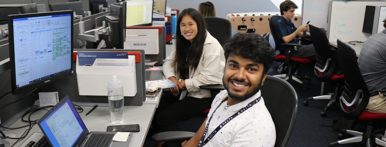 Two students smile for the camera while working on co-op assignments in an office setting in front of large computer screens and desks. 